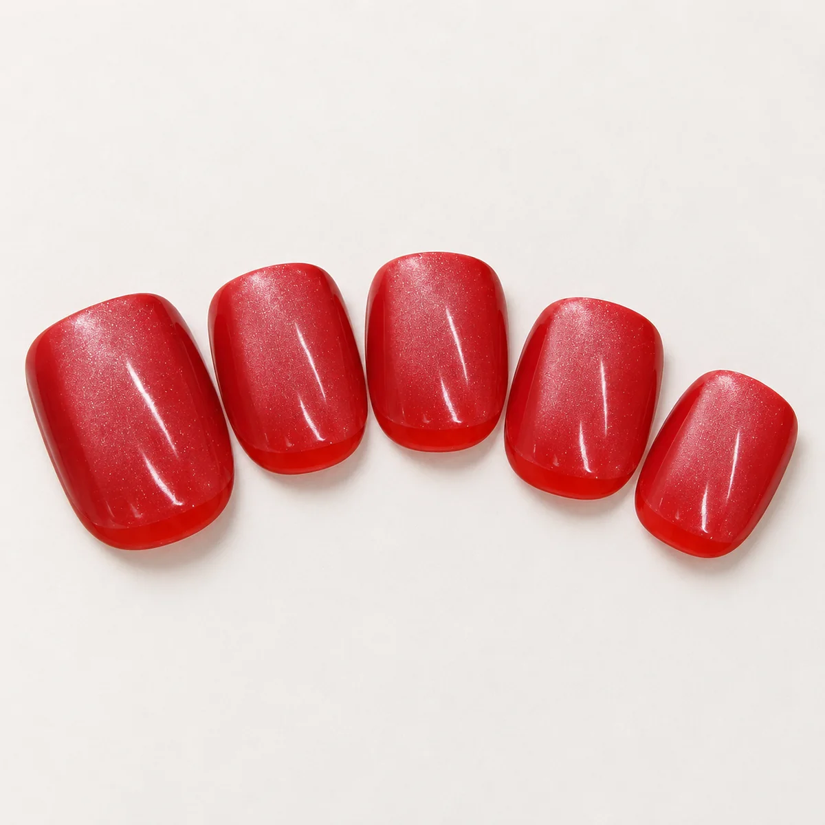 Five selected short rounded-square red press-on nail tips with fine shimmer on an off-white surface.
