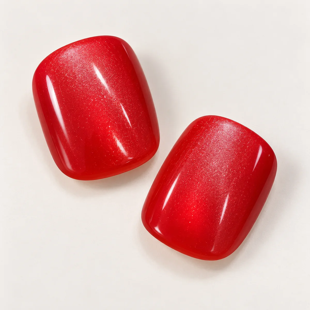 Close-up of two glossy red rounded-square nail tips showing fine reflective shimmer and softly curved corners.